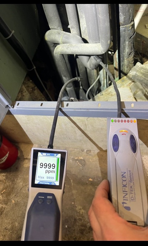 A hand-held electronic refrigerant leak detector held at an insulated line set, its display reading 9999 ppm