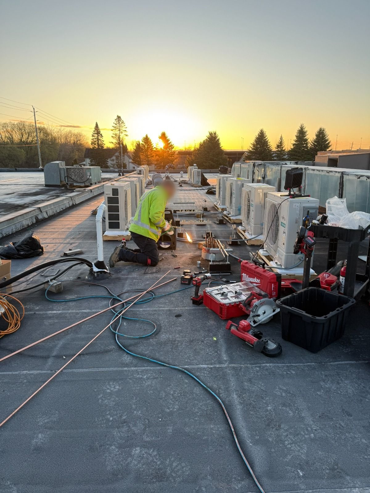 Technician in a high-visibility jacket brazing refrigerant lines on a flat roof at sunset, beside rows of new heat pump units