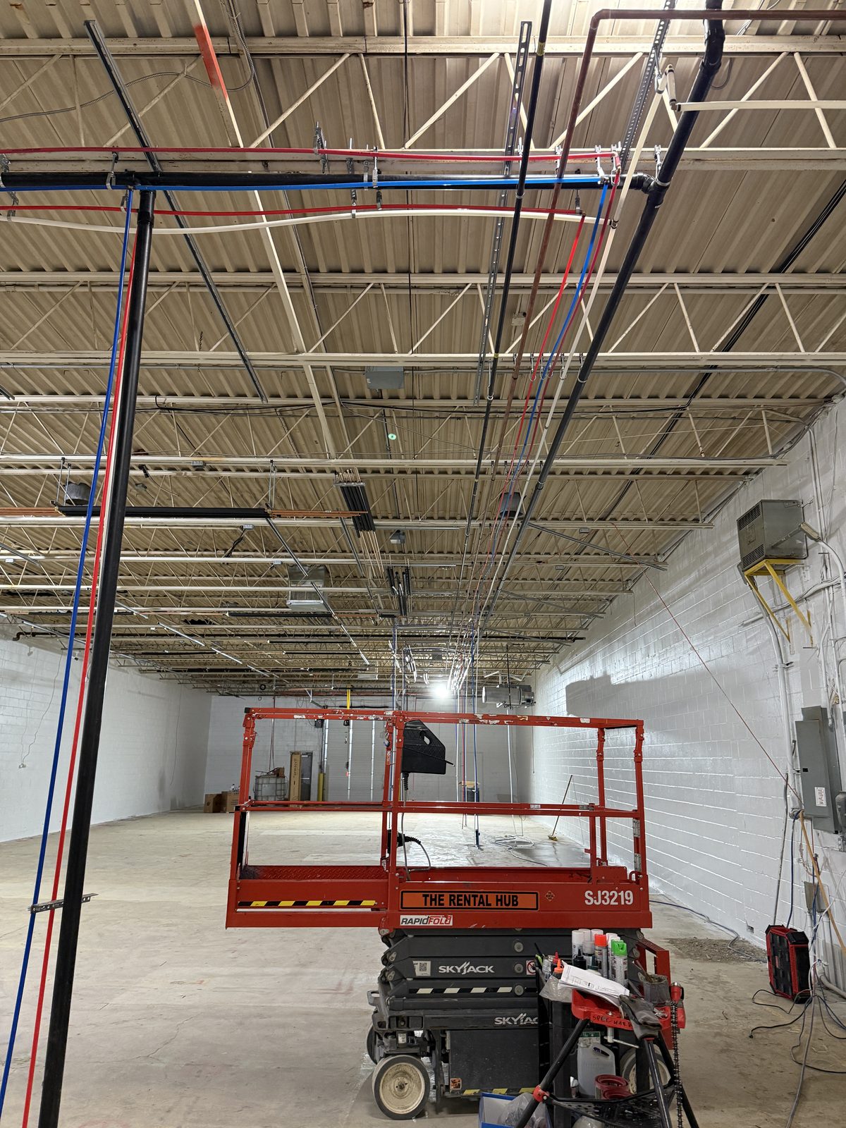 Supply lines run overhead along the warehouse ceiling, with a scissor lift below