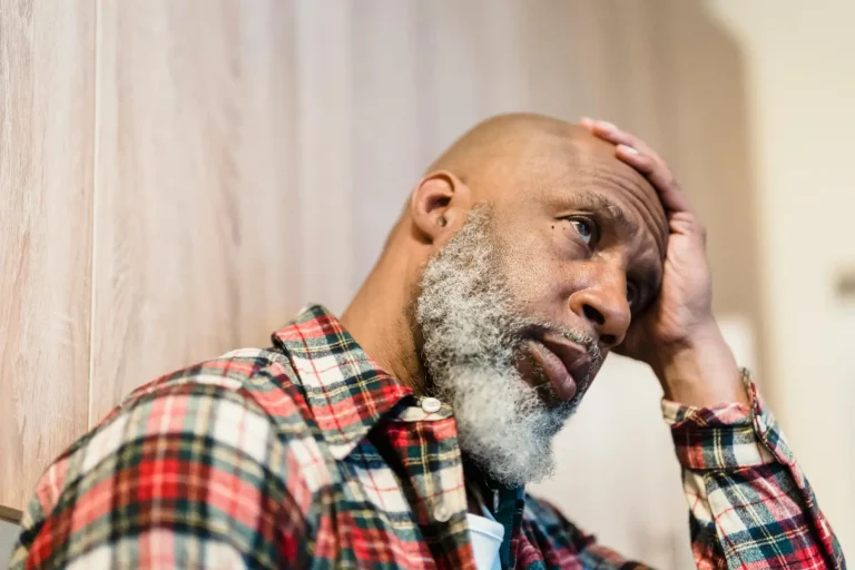 Older man with gray beard in plaid flannel shirt holding his head with a worried expression.