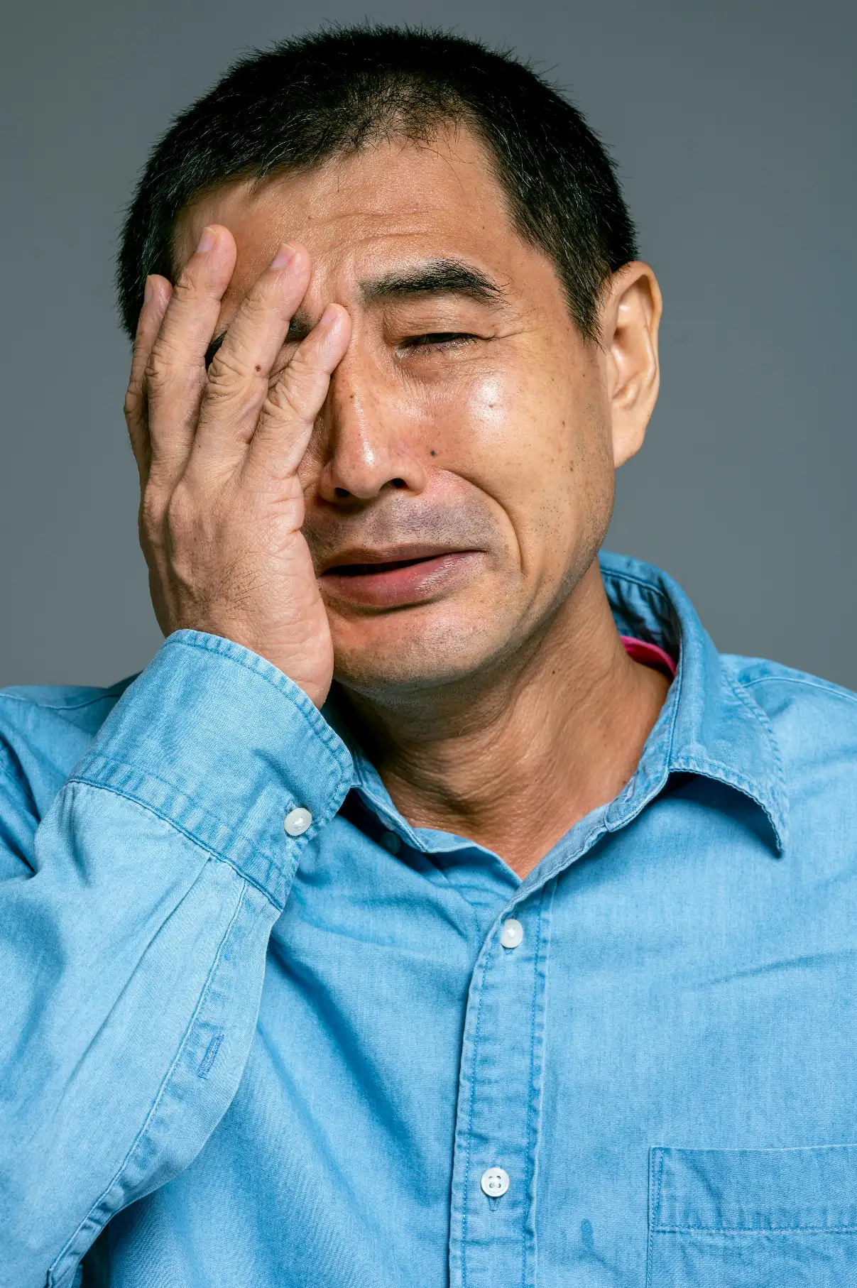 Middle-aged man in a blue cotton shirt covering his face with his hand, looking stressed.